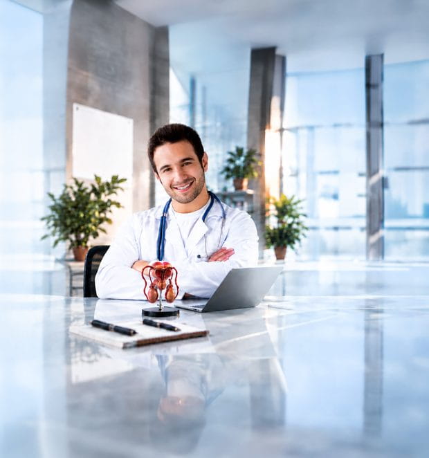 Doctor seated at a clinic desk beside an anatomical model of the male reproductive system